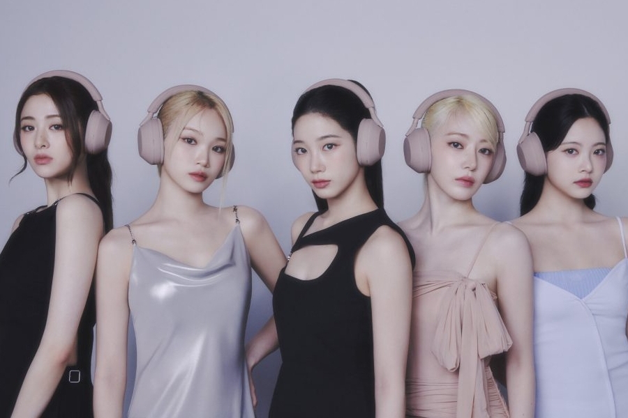 A group of five women, LE SSERAFIM, in stylish dresses and the Sony WH-1000XM5 Smoky Pink Headphones stand against a silver gradient background.