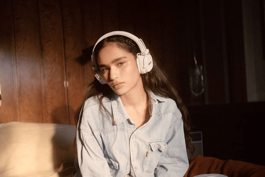 Person wearing the Marshall Major V cream headphones and denim outfit, sitting with legs crossed on a bed in a room with wooden walls.