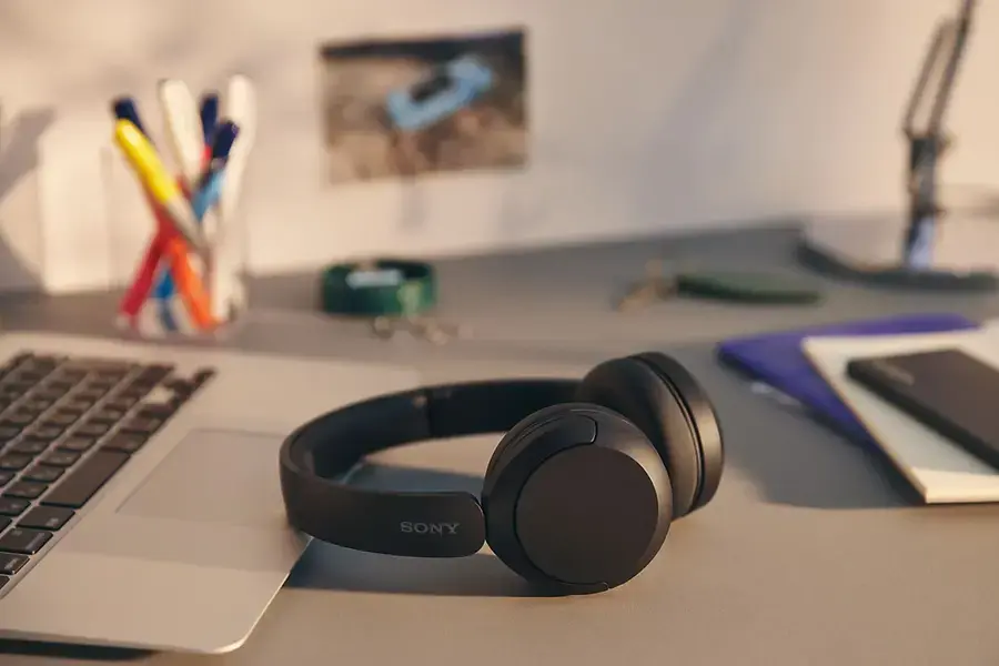 A pair of black Sony wh-ch520 headphones lay on a grey desk that has laptop, mobile phone, and books on it.
