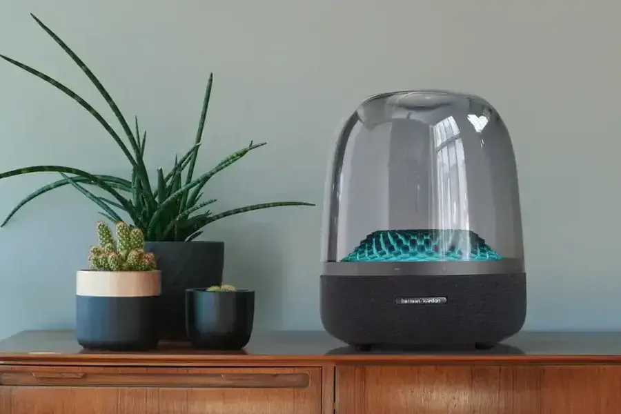 A Harman Kardon Aura Studio 4 speaker on a wooden table. The speaker is a black, round, and glass design with a blue light pattern inside. There are three potted plants on the table, and a wall with a light blue background.